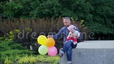 爸爸手里拿着五颜六色的气球，可爱的女儿坐在画城公园的水泥墙上