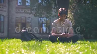英俊的亚洲男人坐在阳光下的草<strong>地上</strong>，<strong>滚动</strong>着他的手机，专注的城市视野，研究着气氛
