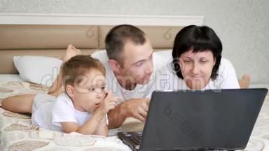 幸福的一家人：爸爸，妈妈和孩子正躺在床上看着笔记本电脑，微笑着。 爸爸亲吻他的妻子和儿子。