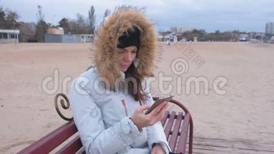 穿着白色<strong>羽绒服</strong>的女人坐在海滩上的长凳上，看着手机上悲伤的视频。