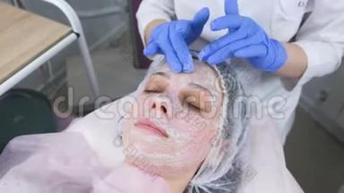 美容师将面膜卷在女人`面部按摩动作上。 美容师面部治疗。