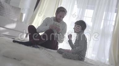 顽皮的胡子爸爸和他的儿子坐在地毯上，拿着钱。 爸爸和孩子把手指放在鼻子上。 快乐