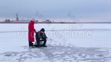 渔夫坐在冰冻的河流上，在冬天的钓竿的帮助下钓鱼，旁边是一位漂亮的年轻女子，看着渔夫