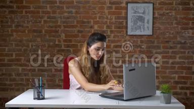 可爱的白种人长卷发女孩坐在桌面上，从她的笔记本电脑上写下笔记，红色的办公室