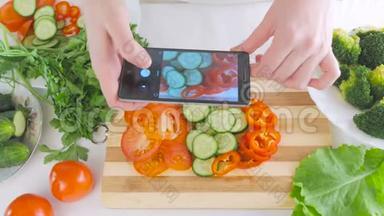 女人`手拿着智能手机，正在为沙拉拍摄新鲜蔬菜的照片，切片在切割板上。
