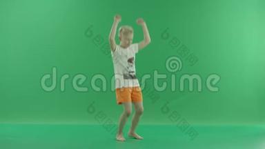 一个穿休闲服装的金发小男孩正在听音乐和跳舞。 他站在绿色屏幕前