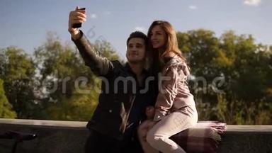 幸福的年轻夫妇在街上用手机自拍，而女孩坐在栏杆上。 漂亮情侣自拍