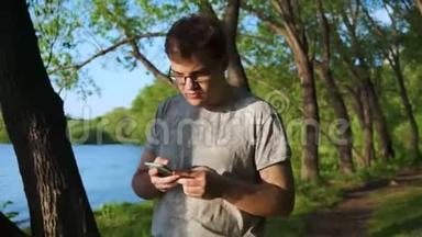 年轻人沉迷于他的手机，在森林里沿着河走，看着他的设备。 库存录像。 男子