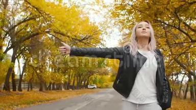 年轻漂亮的女孩走在秋天的跑道上，一个穿着皮夹克和运动服的年轻时髦女人走在沥青路上