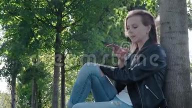 公园里穿着街服的年轻漂亮女孩。 手里拿着智能手机，<strong>坐在树下</strong>的草地上