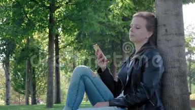 公园里穿着街服的年轻漂亮女孩。 手里拿着智能手机，<strong>坐在树下</strong>的草地上