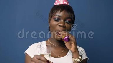 漂亮的非洲女人戴着生日帽，拿着香槟杯，在蓝色背景上吹着生日喇叭。