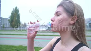 穿着运动衫和运动裤的迷人女孩在体育场喝水美丽的女孩黑发喝水