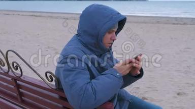 穿着<strong>蓝色羽绒服</strong>的男人坐在沙滩上的长凳上，打着手机，等待着答案。