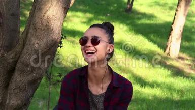 戴着太阳镜的漂亮女孩，积极地笑着，透过社交网络上的新闻，坐在公园的长凳上。