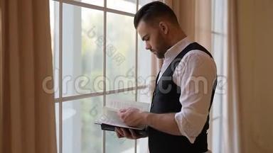 一个年轻人站在窗边翻书。