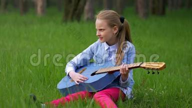 穿着牛仔裤夹克的小女孩玩得很开心，在春天坐在绿草如茵的森林里弹吉他