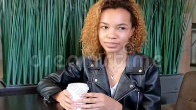 一个身穿<strong>皮夹克</strong>的年轻黑皮肤漂亮的女孩正在绿色的咖啡馆里喝着一杯白色的咖啡
