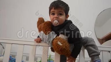 可爱的两岁小孩抱着穿着婴儿身服的玩具熊，打电话给妈妈把他从床上抱下来