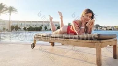 漂亮的年轻女子穿着红色泳衣，一边躺在泳池边的躺椅上，一边用智能手机浏览互联网