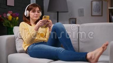 戴耳机的<strong>可爱女孩</strong>躺在客厅的沙发上听音乐和在手机上浏览的画像