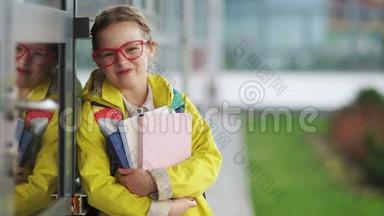 年轻女孩戴着时髦的红眼镜和一件黄色夹克，手里拿着课本，正在学<strong>校门</strong>口微笑