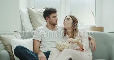 早上看电视的年轻夫妇坐在沙发上，穿着睡衣吃了一大盘爆米花。