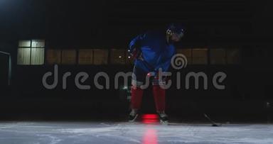 一个曲棍球运动员，手里拿着一根棍子，从黑暗中走出来，把冰球放在冰上
