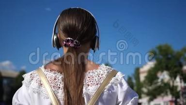 一个年轻的女孩带着耳机走在城市的街道上，听音乐。 穿着白色连衣裙留着长发的少女