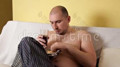 一个又胖又秃又懒的男人吃薯片，喝可乐，坐在沙发上没有外套。 一个卑鄙的人。 有害
