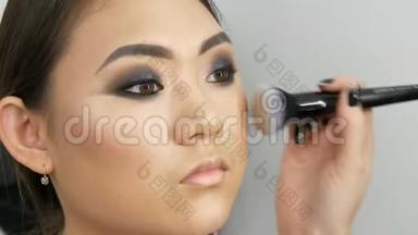 一位专业的化妆师将带有特殊画笔的遮瑕膏应用于视觉工作室特写的亚洲女孩模特脸上