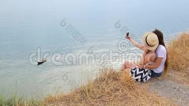 背面观看可爱的积极的金发女孩和黑发女孩坐在悬崖边，并在帮助电话自拍照片