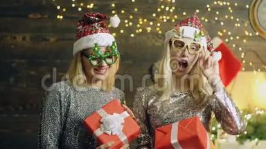 戴着眼镜和帽子的疯狂新年女孩带着礼品盒跳舞。 两个金发女郎穿着银色迷人的礼服和新年