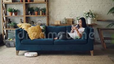 有魅力的亚洲女孩正享受着现代科技的乐趣，她使用智能手机和机器人吸尘器坐在家里的沙发上