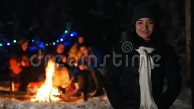 穿着黑色羽绒服的年轻女子站在冬天的森林里。 一群朋友坐在背景上的火炉旁