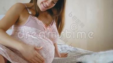 怀孕的女孩和一只猫休息。 一个女人<strong>抚摸</strong>她圆圆的肚子，<strong>抚摸</strong>她的宠物。 床上的动物靠近