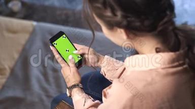 一个棕色头发的漂亮女人，穿着粉色衬衫坐在家里，在她的绿色屏幕上滑动着她的智能手机