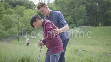 年轻的父亲教他的儿子在公园里骑滑板车。 他支持孩子，帮助他保持平衡