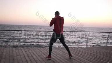 年轻的高个子男子在靠近大海的木地板上做拳击热身运动。 做附加步骤，打孔。 热身运动