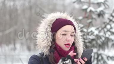 美丽的年轻女孩在冬天的公园里散步时，正在智能手机上浏览社交网络。