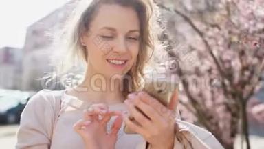 女孩和智能手机。 一个女人在街中央的Skype上聊天。 春天的太阳和风，开花的树