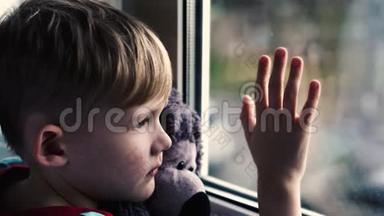 小男孩悲伤地盯着窗外，怀里抱着一只泰迪熊。 坐在窗台上.. 那孩子望着窗外