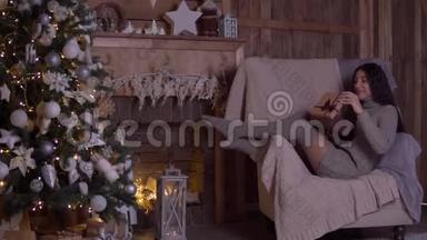 在圣诞节那天，女孩拿着礼物打开盒子，坐在圣诞树附近的椅子上高兴。 高清高清