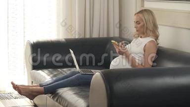一个可爱的怀孕金发女人坐在沙发上，手里拿着一台笔记本电脑和一部手机。 她在寻找