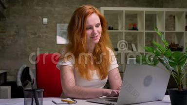 白种人生姜女孩幸福美丽的微笑，坐在她的桌面上，在工作期间使用笔记本电脑，是个自营职业者