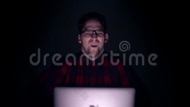 一个男人对着笔记本电脑大喊大叫。 白纸从上面掉下来. 黑暗中监视器后面一个人的肖像。
