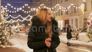 穿着冬季夹克的漂亮女孩站在冬季街道上闪烁的花环的背景上摩擦着她