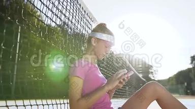 运动女孩坐在网附近的网球场上，使用智能手机。