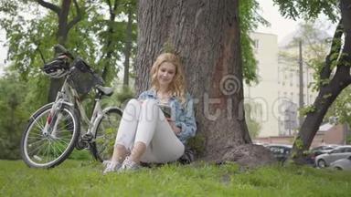 美丽的微笑的女人<strong>坐在</strong>公园的一棵老<strong>树下</strong>读日记。 现代自行车站在附近。 休闲