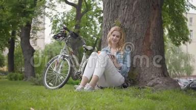 肖像可爱的微笑的女人<strong>坐在</strong>公园的一棵老<strong>树下</strong>读日记。 现代自行车站在附近